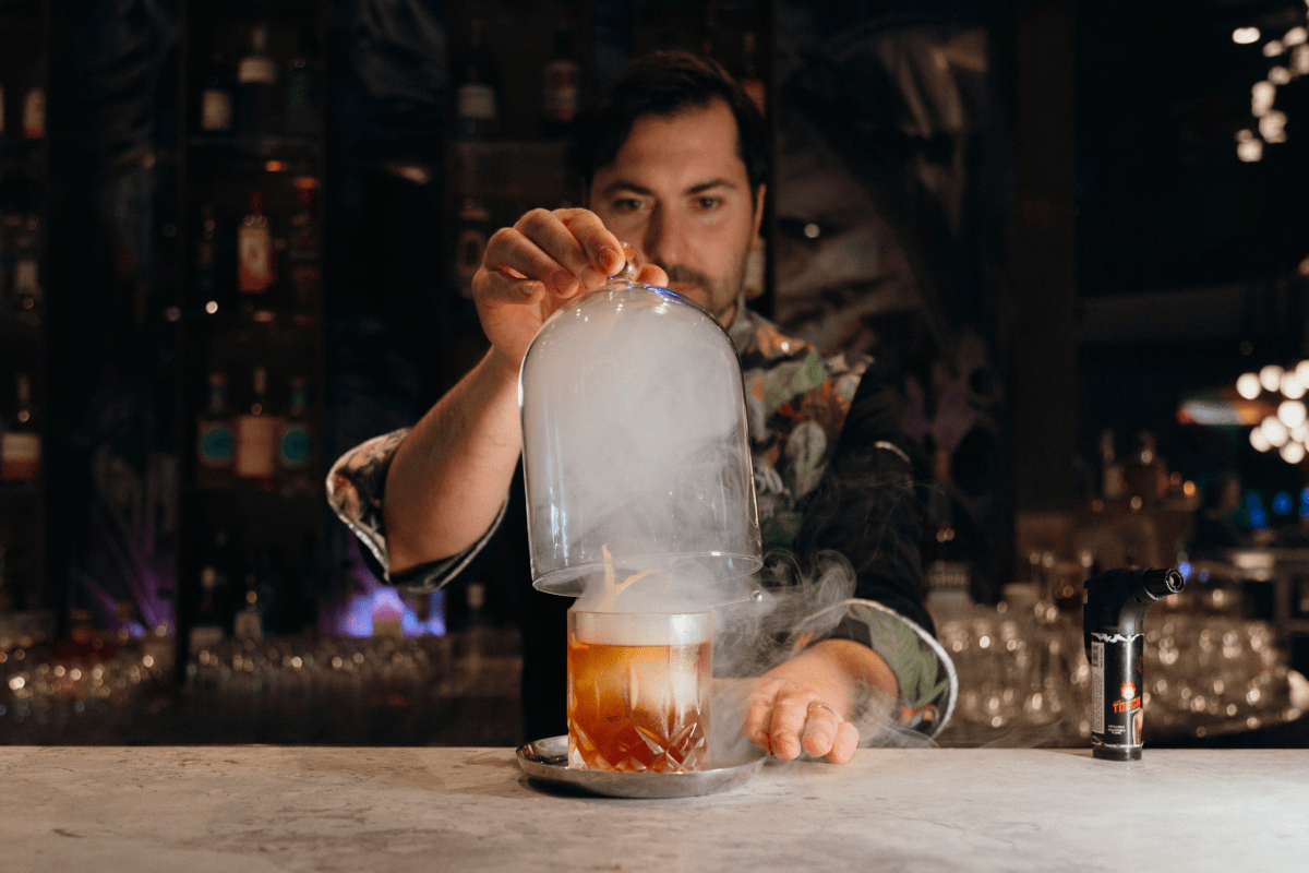 Giacomo Giannotti lifting the glass cover of the Great Gatsy, an Old-Fashioned style cocktail from Paradiso, which uses smoke.